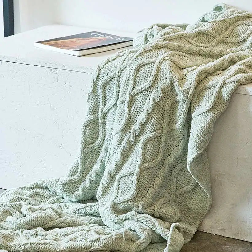Green knitted blanket draped over a white couch with a book on a table.