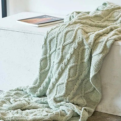 Green knitted blanket draped over a white couch with a book on a table.